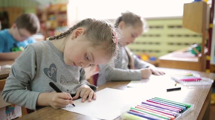 Girls twins sisters draw with felt-tip pens on paper at a table in a classroom. Preschoolers get art education at school. Children draw together