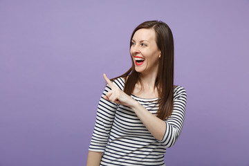 Obraz premium Laughing cheerful young brunette woman in casual striped clothes posing isolated on violet purple background studio portrait. People lifestyle concept. Mock up copy space. Pointing index finger aside.