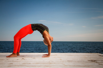 Adult blond woman with short haircut practices yoga on the pier against the background of the sea and blue sky