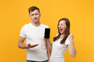 Young couple two friends in white t-shirts posing isolated on yellow orange background. People lifestyle concept. Mock up copy space. Hold mobile phone with blank empty screen, doing winner gesture.