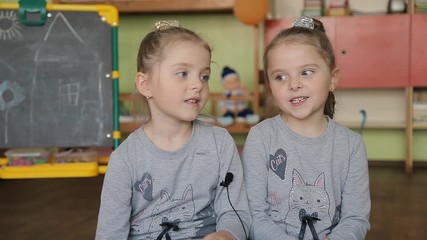 Twin girls sitting on a chair answers questions. Girls give interviews in a classroom against the background of the blackboard. Themes sister siblings twins affection