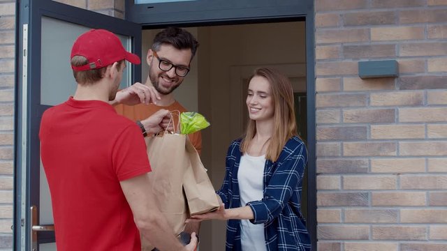 Food delivery man service giving fresh supermarket food package to the customers. View of young pretty couple smiling taking order paying with paypass technology smartphone.
