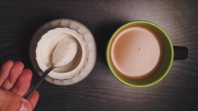 Overhead slow motion of pouring sugar from a bowl in the ceramic cup with black coffee with milk by a caucasian man on wooden table