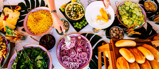 Aerial vertical top view of table full of mixed food for party catering celebration people concept - hands serving and. taking on dish - coloured background with healthy ingredients