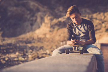 teenager at the beach sitting on a wall using his phones with his skateboard - boy wearing jeans focused on the social or playing video games