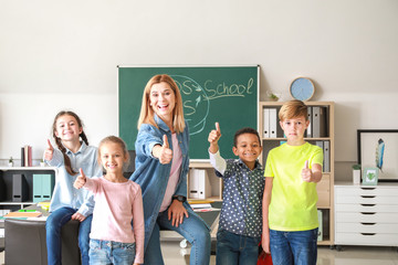 Cute children with teacher showing thumb-up gesture in classroom - obrazy, fototapety, plakaty