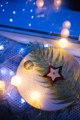 Glitter shiny branch with leaves with Christmas decoration on plate near window in the evening with warm garland lights.