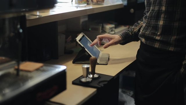 Male cashier hands are using a mobile digital tablet behind a coffee shop counter. A close-up