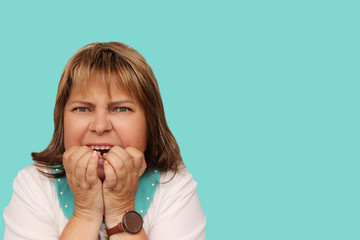 pretty middle-aged woman with a grimace of stress and despair bites clenched hands, turquoise background, isolate, close-up, copy space