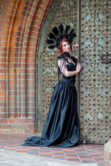 Fototapeta na wymiar Gothic Girl in Long Black Dress. Wearing Artistic Feather Crown with Roses. Posing Against Old Castle Gates.