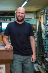 Portrait of a shoemaker bald man with beard, posing standing at his workshop.