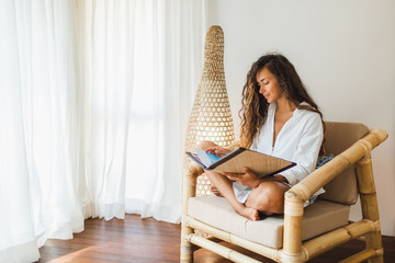 Young brunette woman sitting in wooden bamboo chair indoors and reading book or magazine. Eco style...
