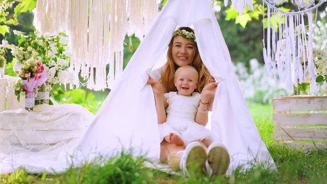 A baby with her mother hides in the wigvam on the green grass. Outside shooting, the garden on the background. Portrait. Party atmosphere.