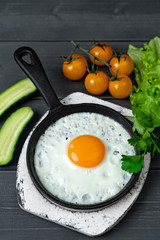 fried eggs in cast iron frying pan on white wooden stand with yellow cherry tomatoes, and cucumbers. Tasty, healthy breakfast.