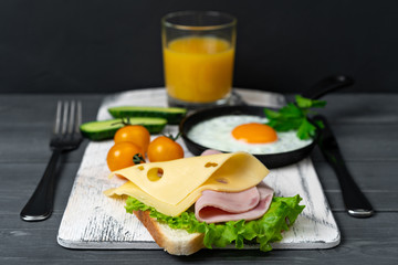 Breakfast of toasts with cheese, ham, lettuce, scrambled eggs in pan, cherry tomatoes, cucumbers and orange juice. Serve on cutting board. Nutritious breakfast.