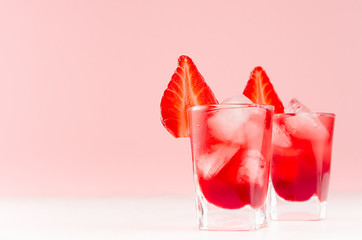 Cold red alcoholic liquors with ice cubes, strawberry slice in two shot glasses on white wood table and pastel pink wall, copy space.