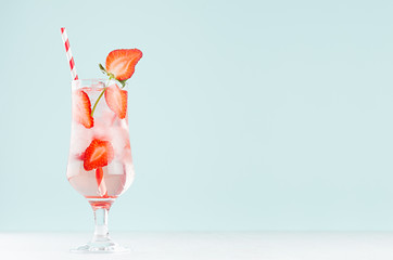Cold strawberry cocktail with ripe berry pieces, ice cubes, tonic, straw in light soft pastel blue color background on white wood board.