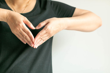 A woman use hands display love symbol by splice fingertips together to be heart shape at front of left chest.