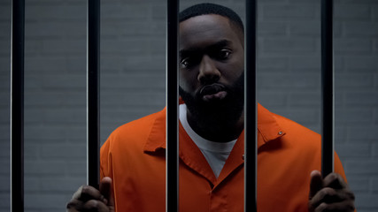 Black male prisoner waiting for sentence in prison cell, looking sadly to camera