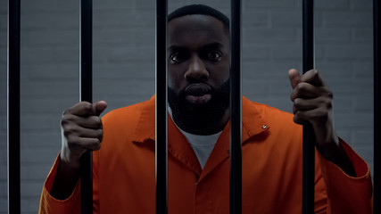 Terrified afro-american prisoner holding bars, looking at camera with fear