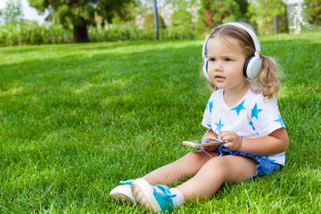 Little cute girl sitting in the Park on the grass on a Sunny summer day and listening to music in white headphones. Lifestyle, happy summertime, summer outdoor portrait