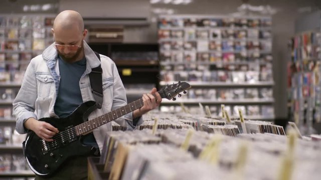Trucking Medium Shot Of Bearded Caucasian Male Musician With Shaved Head, Wearing Denim Jacket And Tinted Sunglasses, Standing In Aisle Of Specialty Music Store And Trying Out Electric Rock Guitar