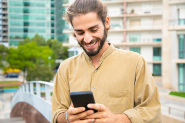Happy excited hipster guy holding smartphone and smiling at screen. Young man in casual standing in city, using mobile phone, texting message, getting good news. Good news concept