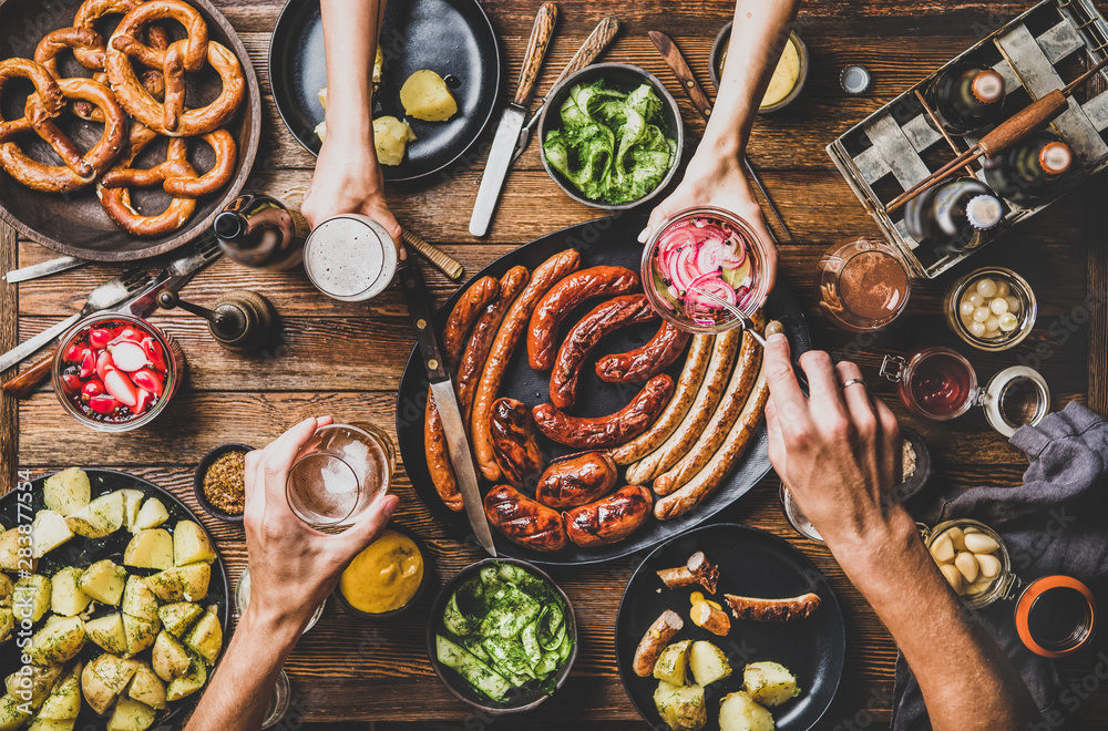 Wall mural Flat-lay of Octoberfest party dinner table with grilled meat sausages, German pretzel pastry, potatoes, cucumber salad, sauces, beers and peoples hands with food over dark wooden background, top view