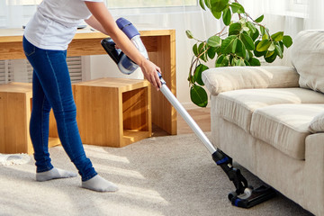 Portrait of young woman in white shirt and jeans cleaning carpet under sofa with vacuum cleaner in...