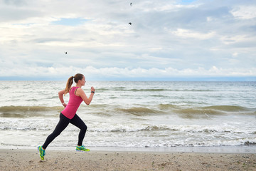 Young fitness woman runner running at seaside, copy space. Girl working out on beach at summer morning, full length portrait. Side view. Healthy lifestyle concept