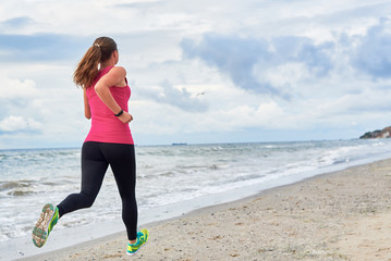 Back view of young fitness woman runner running at seaside, copy space. Girl working out on beach at summer morning, full length portrait. Healthy lifestyle concept