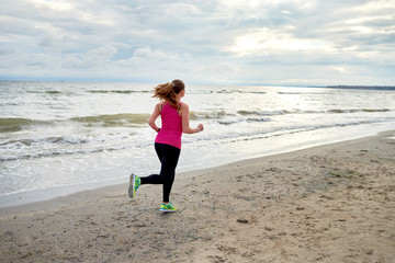 Back view of young fitness woman runner running at seaside, copy space. Girl working out on beach at summer morning, full length portrait. Healthy lifestyle concept