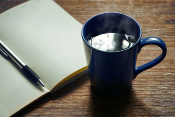 Hot coffee in blue cup with diary book on wood table.
