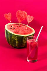 Useful watermelon juice with a tube and a large ripe watermelon decorated with hearts on sticks on a red background. Concept of summer mood, harvest of seasonal berries and fruits