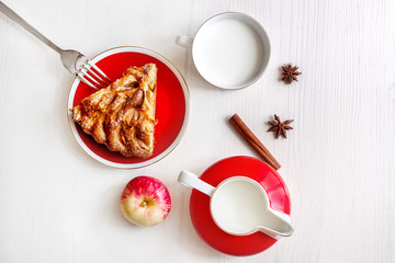 A piece of apple cake with cinnamon, fresh apple, milk cup and jug on the light wooden table. Top view