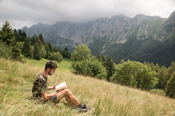 Caucasian man sitting in the grass and reading a book in a mountain meadow. Beautiful natural landscape behind. Human and nature connection concept