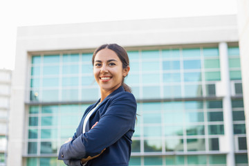 Fototapeta premium Happy successful young businesswoman posing outside. Beautiful Latin woman wearing formal jacket, standing near office building, looking at camera and smiling. Successful career concept