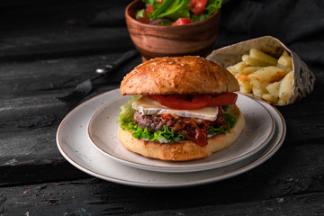homemade burger on a rustic plate, on a dark wooden table, dark background, French fries and the wooden bowl of salad