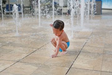 Adorable little Asian baby boy having fun on water stream of a sprinkler. Kid playing in playground fountain in aqua park.