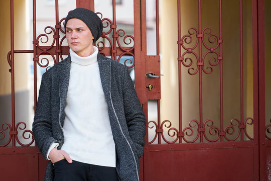 Stylish hipster man wearing elegant fashionable grey coat, stylish white sweater, black jeans near openwork fencel. Autumn collection of fashionable men's clothing.