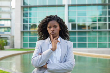Fototapeta na wymiar Serious pensive female manager posing outside. Young black woman wearing formal suit, standing near office building, touching chin and looking at camera. Serious professional concept