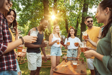 Group of happy friends having beer and barbecue party at sunny day. Resting together outdoor in a forest glade or backyard. Celebrating and relaxing, laughting. Summer lifestyle, friendship concept.