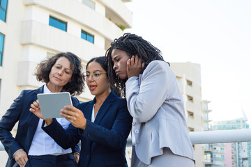 Focused female colleague with tablet pc. Low angle view of professional multiethnic businesswomen standing together on street and using tablet computer. Technology concept