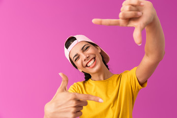 Image of cheery joyful woman wearing cap laughing and showing photo frame with fingers