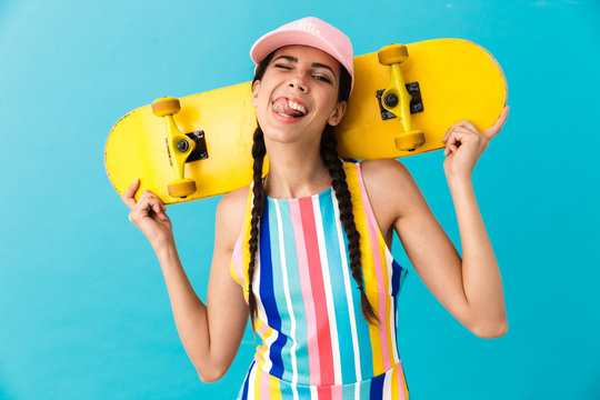 Image of joyful caucasian woman wearing cap winking while carrying yellow skateboard on her shoulders