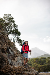 Naklejka na ściany i meble Slim fit woman in sportwear standing with hiking backpack and sticks on the rock and looking up