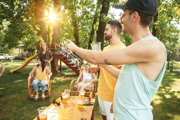 Group of happy friends having beer and barbecue party at sunny day. Resting together outdoor in a forest glade or backyard. Celebrating and relaxing, laughting. Summer lifestyle, friendship concept.