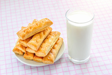 Glass of cow milk and sweet puff pastry cookies on a white saucer over white pink checkered tablecloth. Tasty breakfast with milk.