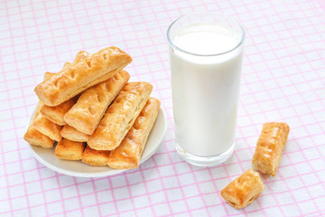 Sweet puff pastry cookies on a white saucer and glass of cow milk over white pink checkered tablecloth. Tasty breakfast with milk.