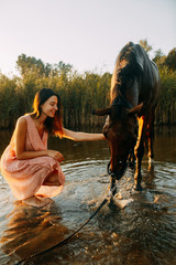 Woman sits next to the horse that drinks water at sunset.